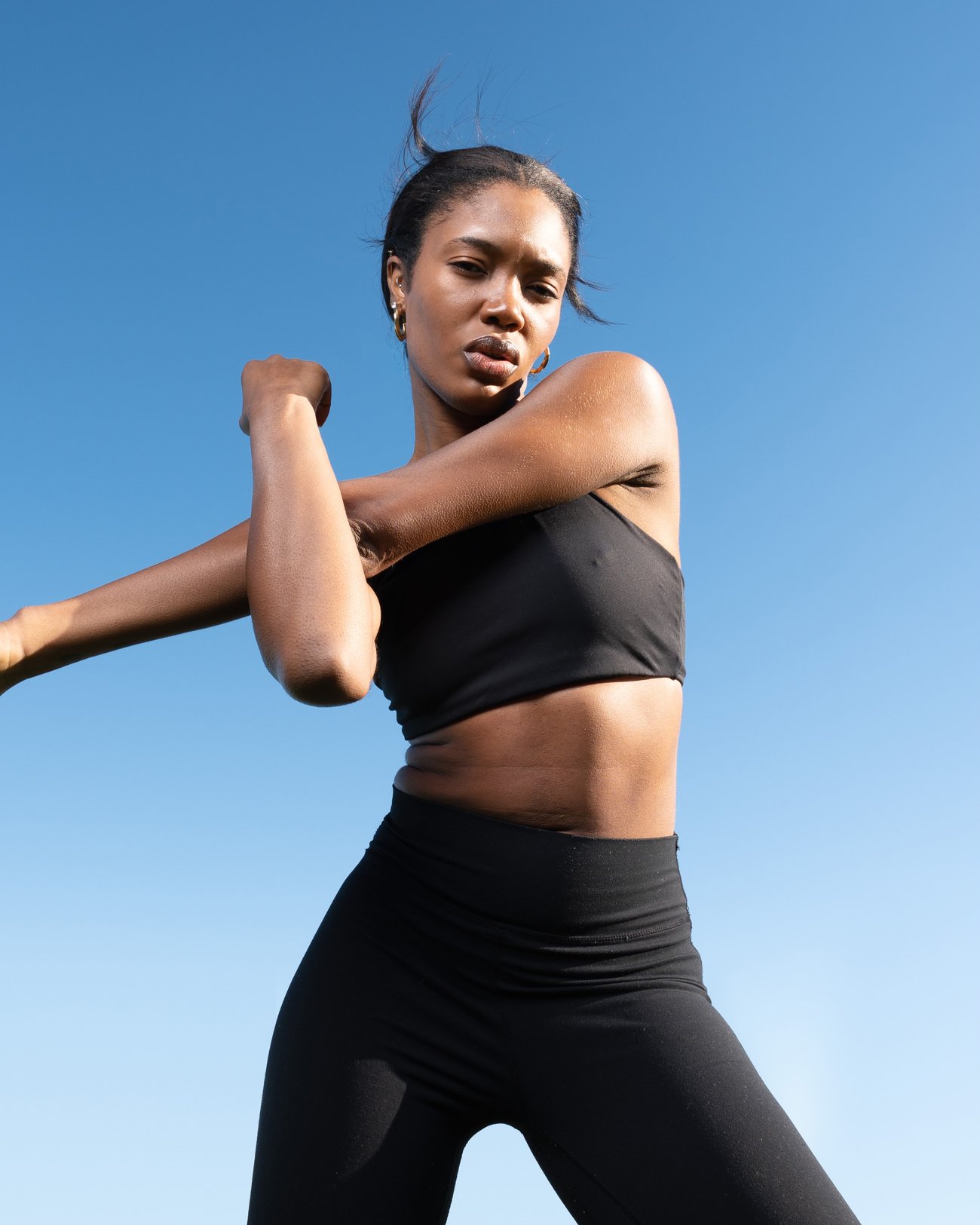 Ruqayyah in a standing cross-body shoulder stretch against a clear sky, the arm and shoulder held in one straight line