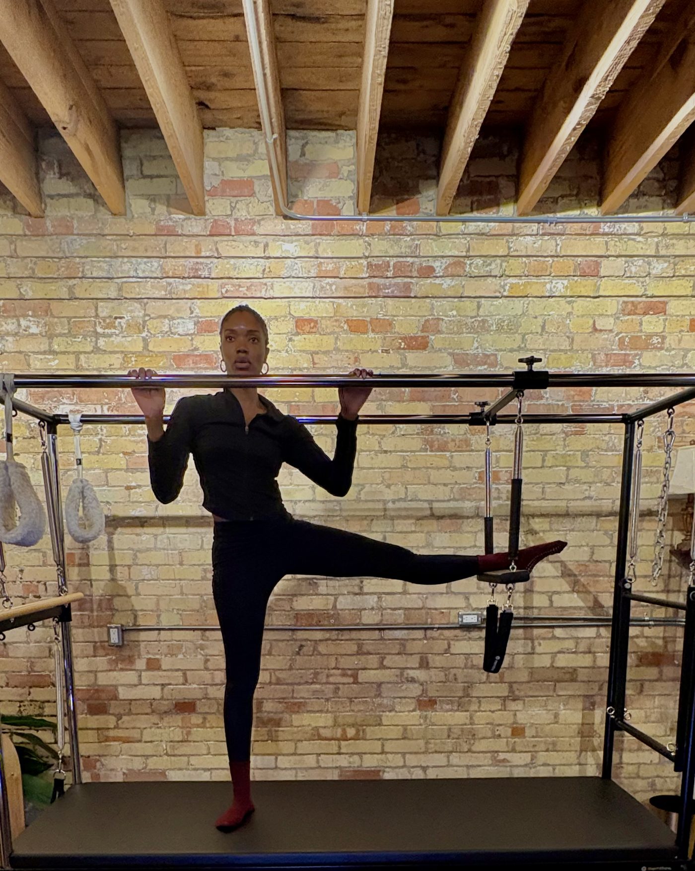 Ruqayyah at the Cadillac trapeze table in her downtown Toronto studio, standing on the table with one leg extended to the side and both hands on the push-through bar