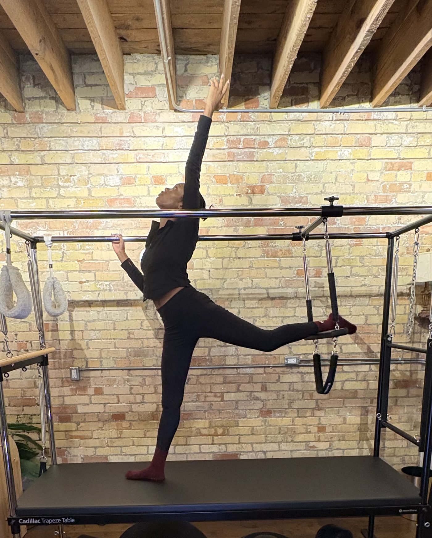 Ruqayyah standing on the Cadillac table, one arm reaching straight overhead and the opposite leg extended, showing a long line from the supporting foot to the fingertips